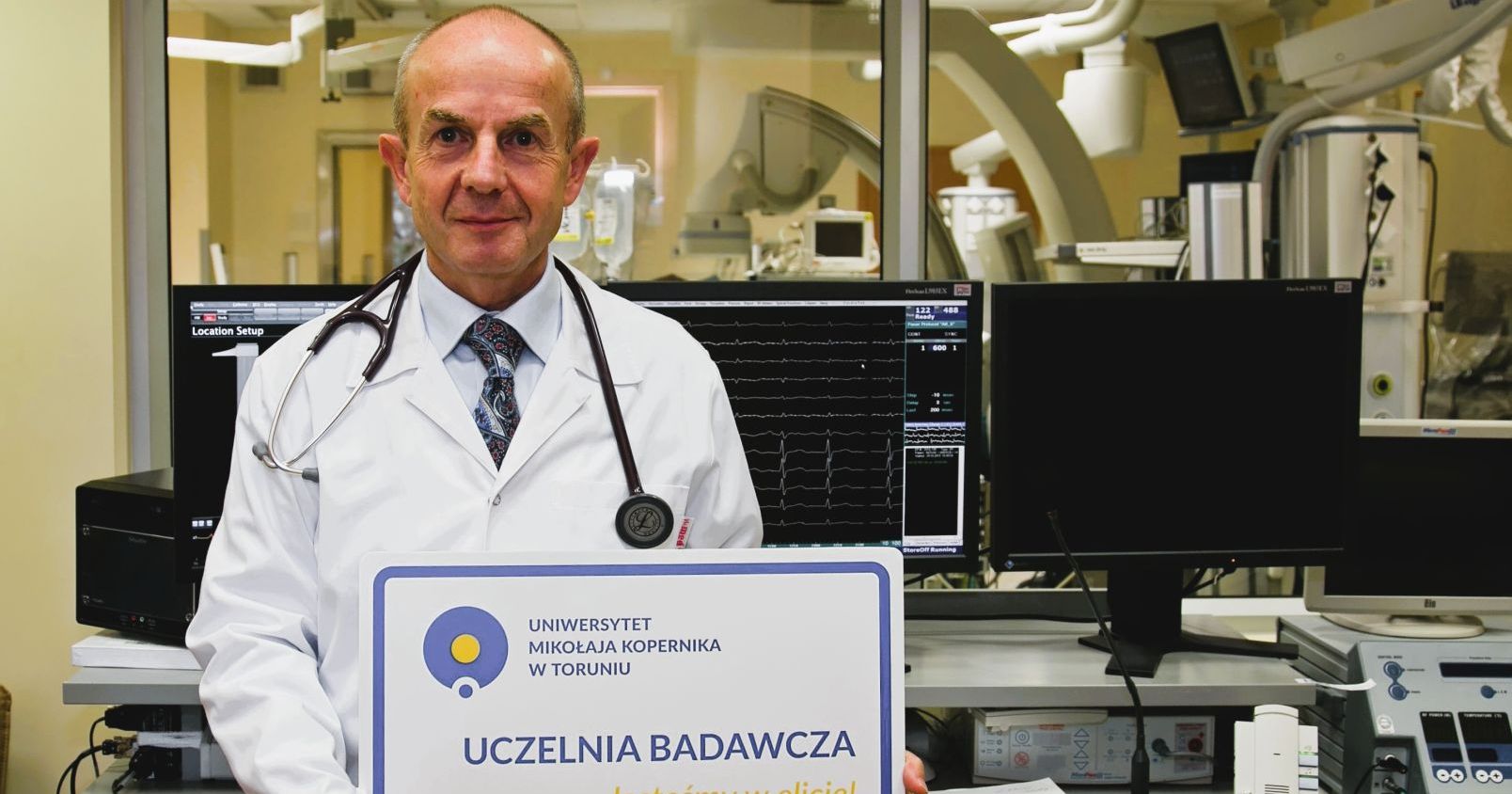 Prof. dr hab. Jacek Kubica z Wydziału Lekarskiego Collegium Medicum UMKe Prof. dr hab. Jacek Kubica z Wydziału Lekarskiego stoi w fartuchu ze stetoskopem w laboratorium z tabliczką uczelnia badawcza