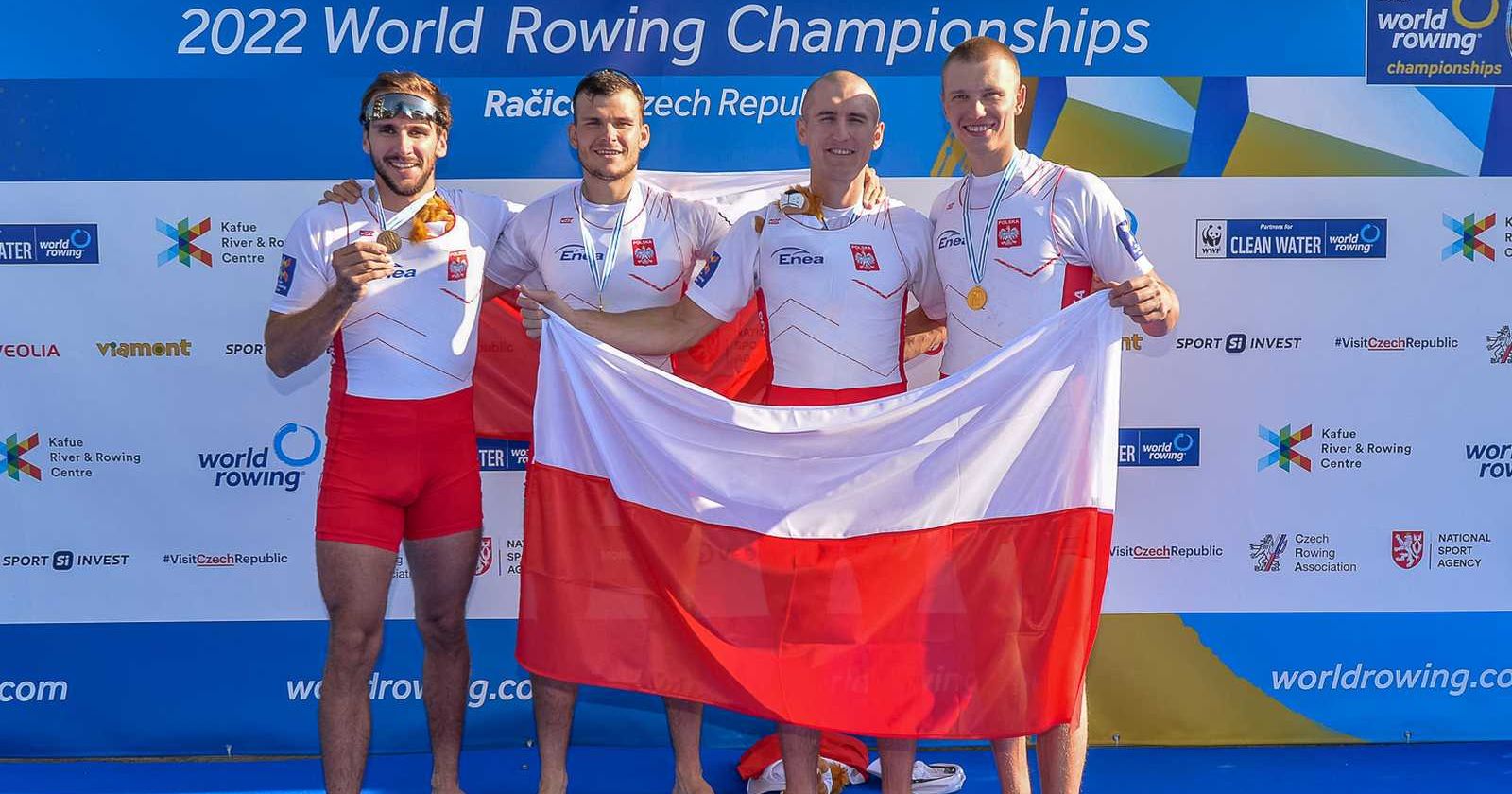 Złoci medaliści mistrzostw świata 2022 Reprezentanci Polski ze złotymi medalami i flagą Polski pozują na ściance z napisem "2022 World Rowing Championships"