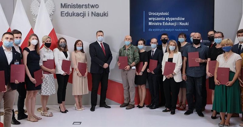  Zdjęcie przedstawia grupę laureatów nagrody. Niekótrzy trzymają w rękach teczki. W tle widać trzy flagi polski i napisy: Ministerstwo Edukacji i Nauki oraz Uroczystość wręczenia stypendiów Ministra Edukacji i Nauki dla wybitnych młodych naukowców