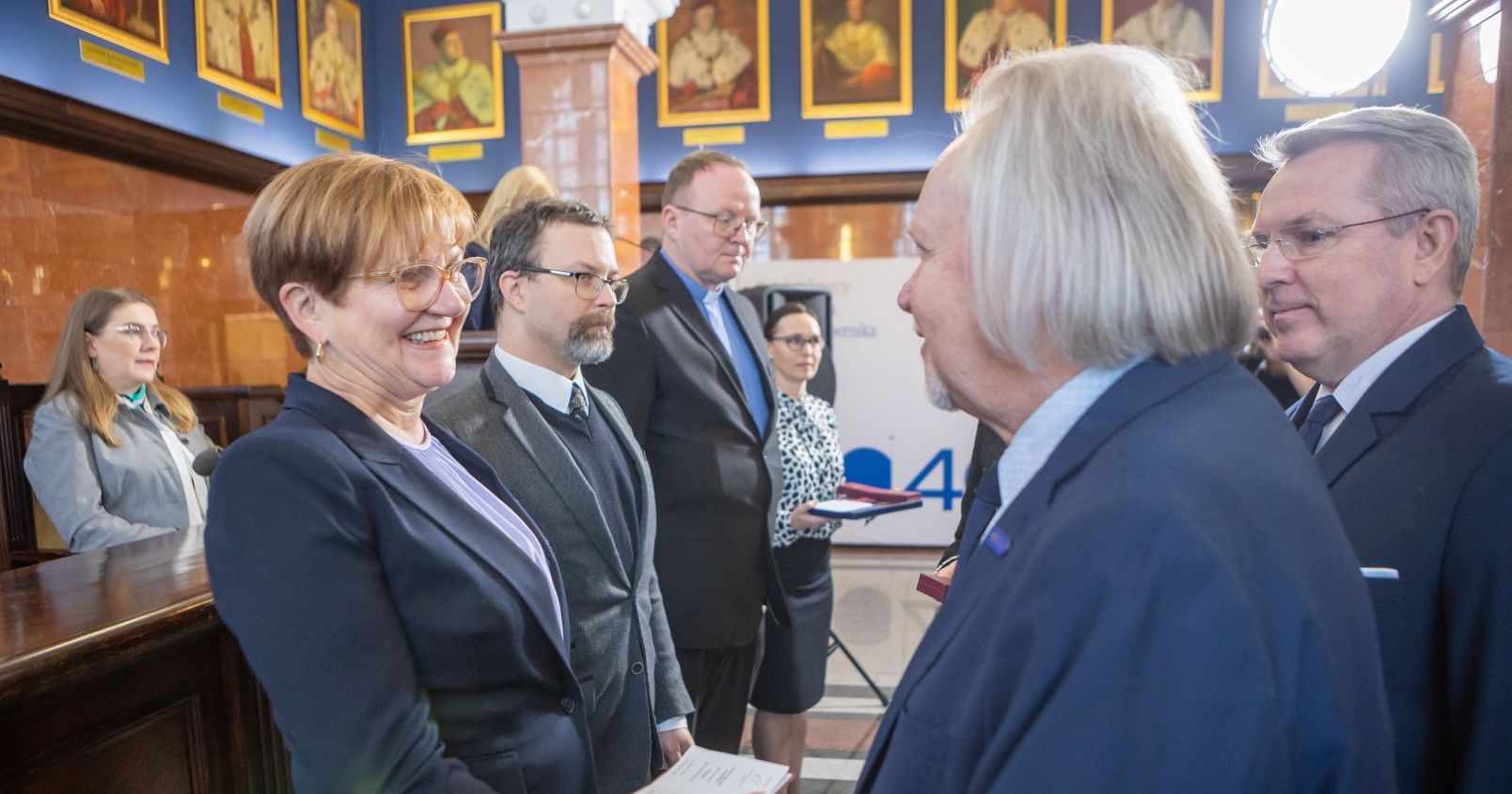 Pracowniczki i pracownicy UMK odebrali odznaczenia z rąk rektora prof. Andrzeja Tretyna, wicewojewody kujawsko-pomorskiego Piotra Hammerlinga i kujawsko-pomorskiej kurator oświaty Grażyny Dziedzic Rektor wręcza odznaczenie prof. Magdalenie Osińskiej. Obok rektora wicewojewoda, w kolejce stooją następne osoby, które otrzymają medal. w tle portrety rektorów UMK w reprezentacyjnej sali senatu Collegium Maximum