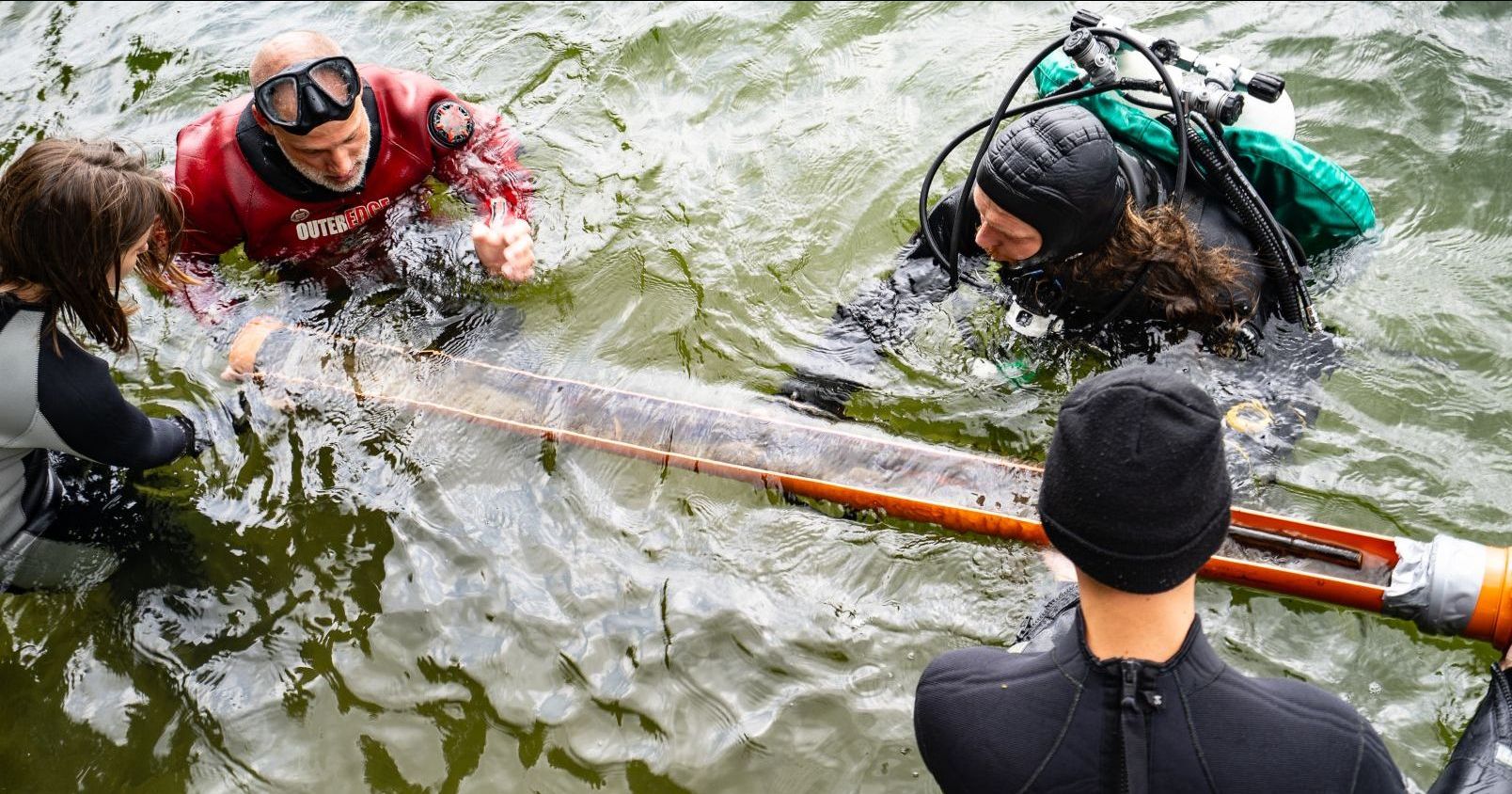 Four divers lift an orange tube with a spear on a pole from the lake