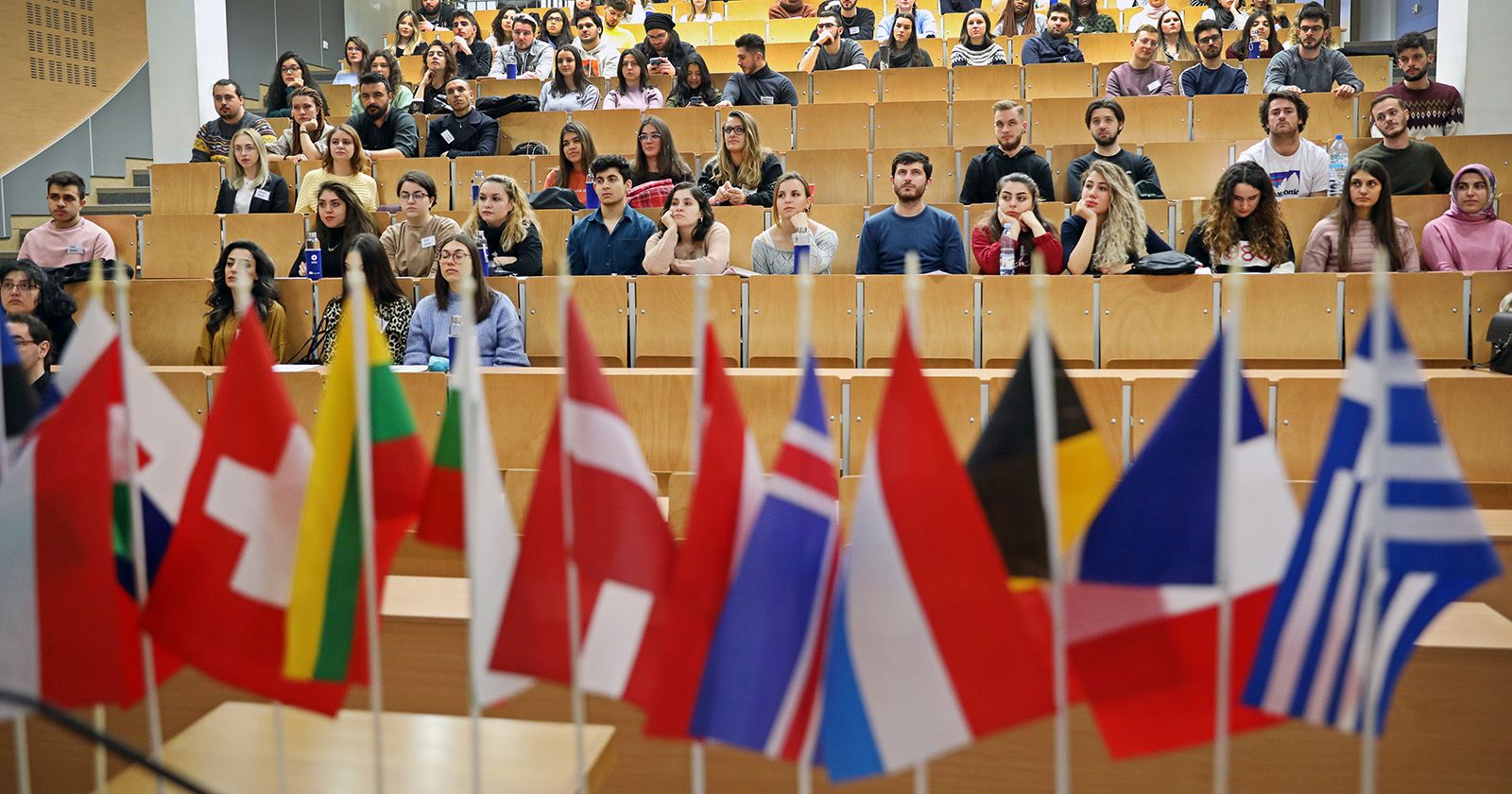  Grupa osób siedzi w audytorium na zajęciach/wykładzie, na pierwszym planie małe flagi europejskich państw
